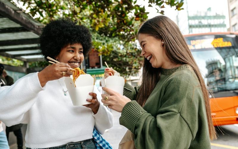Two women laughing and eating street food in the city.